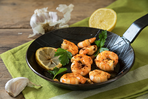 Fried king tiger prawns with garlic, lemon and italian parsley in a black iron pan on a green napkin and rustic wood, selected focus