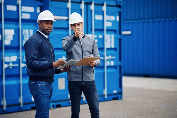Two engineers tracking inventory using a clipboard and digital tablet while standing by freight containers on a large commercial shipping dock