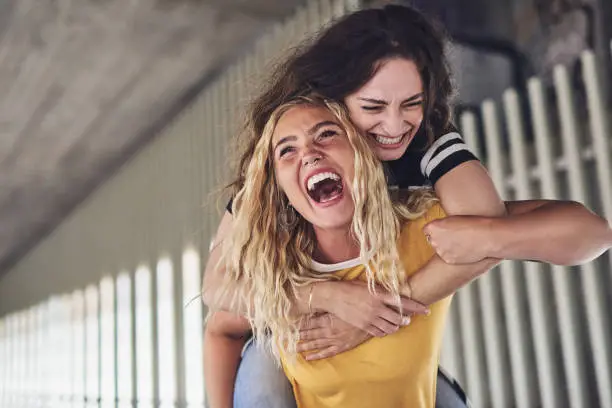 Laughing young woman carrying her girlfriend on her back while having a fun night out together in the city
