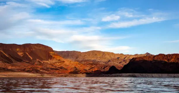 Sunset view of Lake Mead hills, mesas and mountains in tones of brown, orange, purple, mauve and red with water reflections, blue sky and clouds.