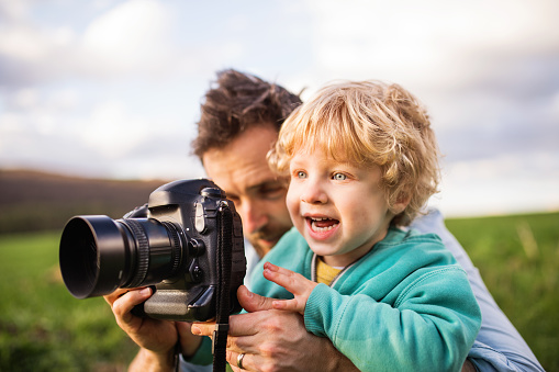 A father and his toddler son with a camera outside in green sunny spring nature.