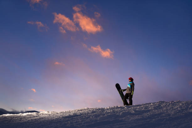 Girl snowboarder stands on a hillside against  dark sunset sky Girl snowboarder stands on a hillside against the dark sunset sky ski resort flash stock pictures, royalty-free photos & images