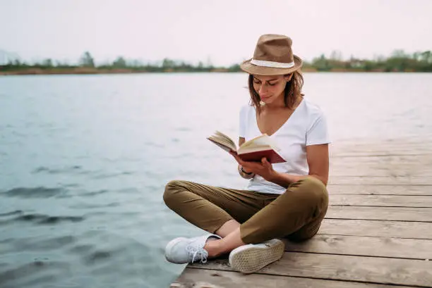 Photo of Portrait of a girl reading a book while sitting on a small wooden wharf.