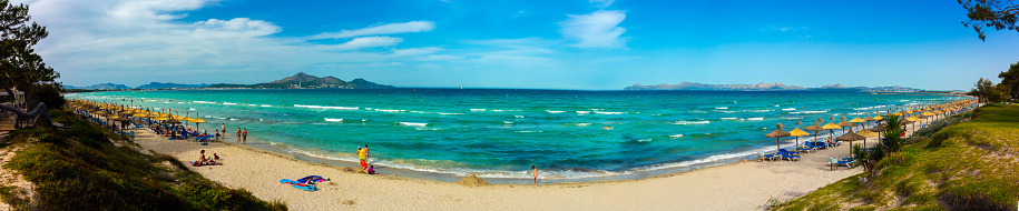 Alcudia, Mallorca, Spain - July 07, 2017: Row of beach umbrellas on the sandy beach and clean emerald green sea. Panoramic image. People in the background.