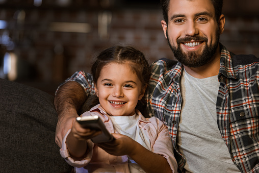 happy father with daughter sitting in couch, hugging and watching TV at home