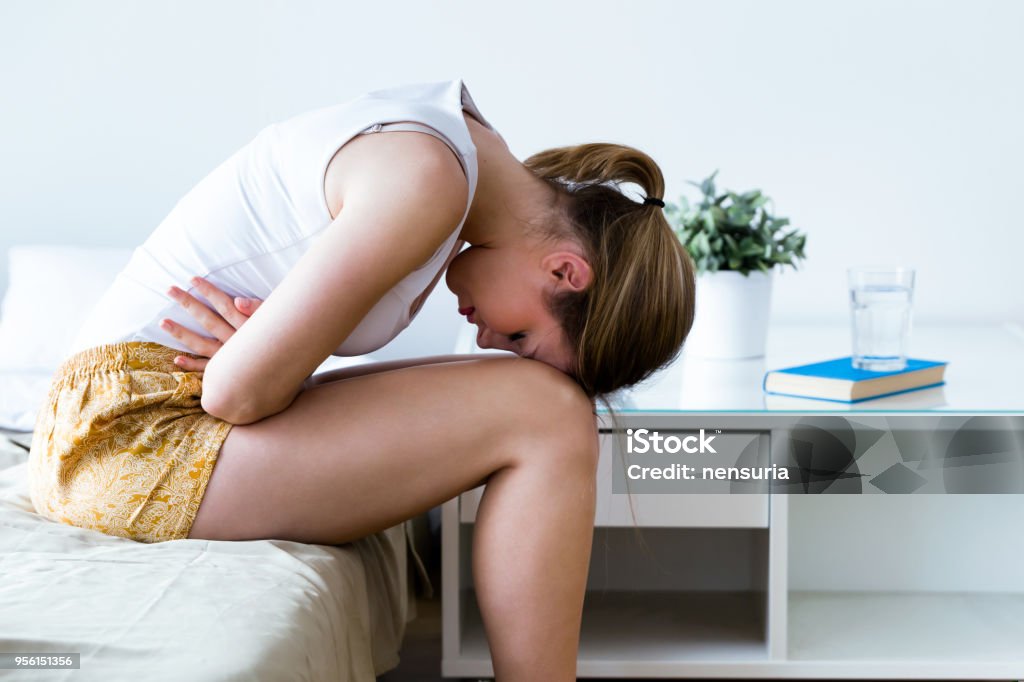 Unhealthy young woman with stomachache leaning on the bed at home. Shot of unhealthy young woman with stomachache leaning on the bed at home. Pain Stock Photo