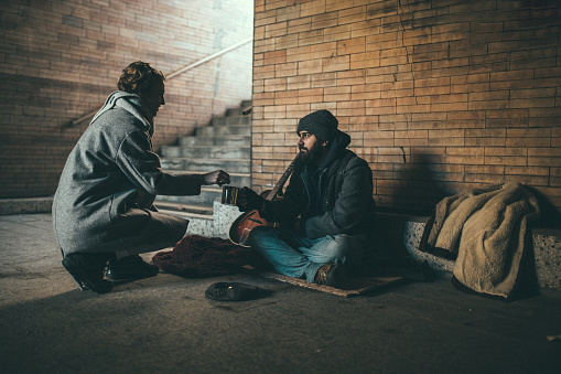 One man, young homeless sitting on the street and begging, woman handing him some money.