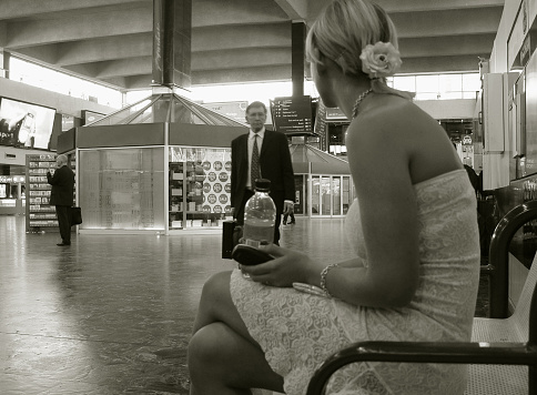 Scene at the Euston train station (London, UK): young woman is waiting for someone, she is sitting, holding her mobile phone and looking towards smartly dressed older man with briefcase walking in her direction and looking in tern on her. Another smartly dressed man with briefcase standing not far, looking in other direction; display advertising showing advert of the mobile phone.