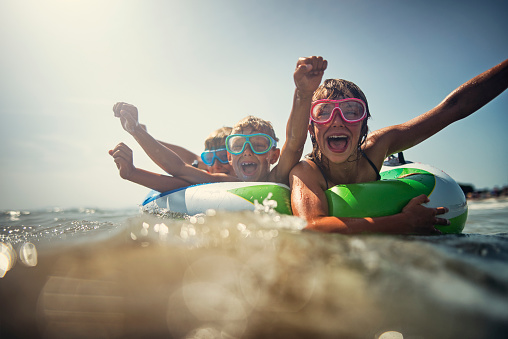 Kids are splashing and having fun on swim ring floating on the sea. Kids are cheering at the camera.\nNikon D810