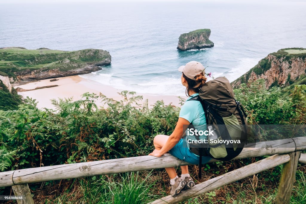 Menina viajante senta-se na cerca de madeira e fica na praia do oceano - Foto de stock de Longa Caminhada royalty-free