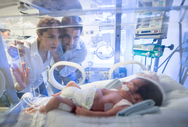 Worried young couple looking at their premature newborn in an incubator with oxygen at neonatal intensive care unit Worried young couple looking at their premature newborn in an incubator with oxygen at neonatal intensive care unit and nurse standing in the background incubator stock pictures, royalty-free photos & images
