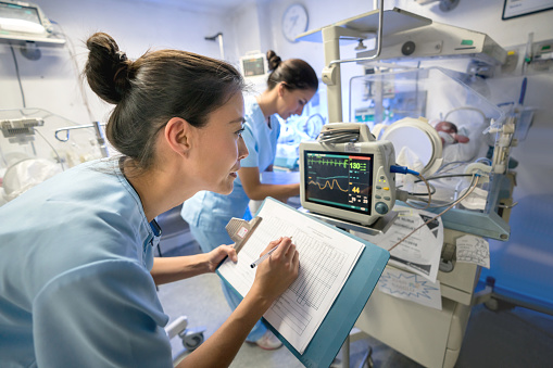 Team of nurses in neonatal intensive care unit checking on a premature's vital signs and taking notes writing the numbers on a clipboard and the other one completing the medical chart