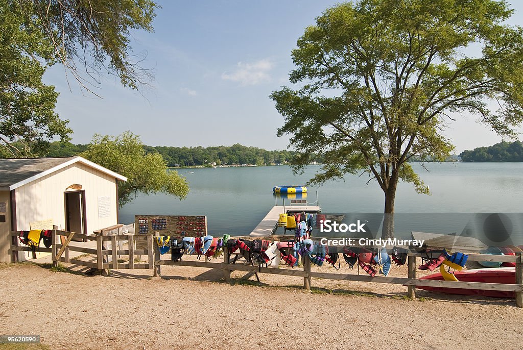 Summer Camp Lake with Life Jackets Hanging on Fence View of beach at Summer Camp with boat house, changing room, wooden fence with life jackets and life preservers hanging, trees, lake, boats. Summer Camp Stock Photo