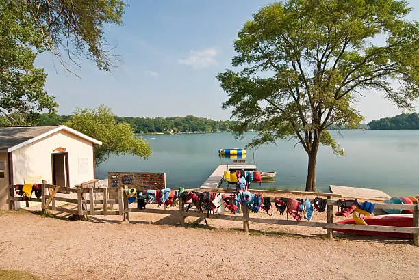View of beach at Summer Camp with boat house, changing room, wooden fence with life jackets and life preservers hanging, trees, lake, boats.