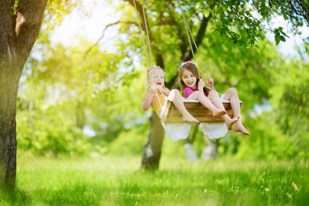 dos lindas hermanitas divirtiéndose en un columpio en el jardín de verano hermoso en día cálido y soleado al aire libre - child swing swinging spring fotografías e imágenes de stock