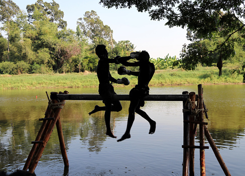 Khai Bang Rachan, Sing Buri, Thailand April 14, 2018: Silhouette or the dark shape and outline of two man boxing on the timber over the water, It is traditional water Thai boxing (or Muay Talay) ancient Thai fight above the water. on Songkran Festival at Pho Kao Ton temple.