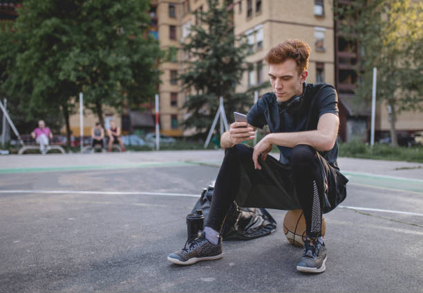 istock Hipster sitting on a basketball texting 956318526