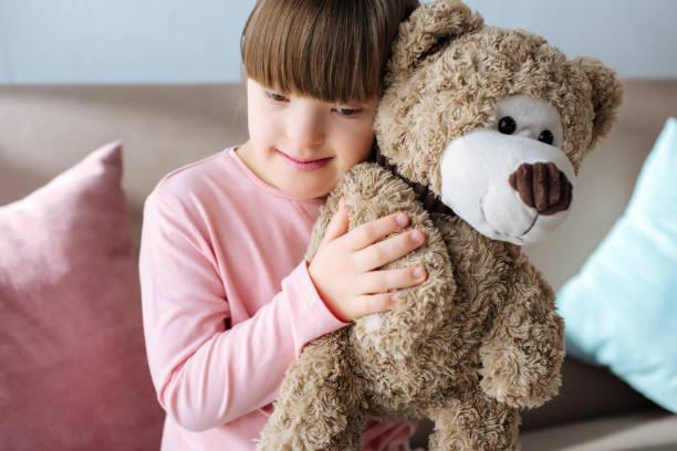 istock Kid with down syndrome sitting on sofa and hugging teddy bear 956321280