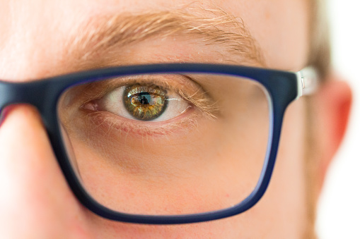 Macro extreme close up depicting a male human face and one eye wearing a trendy pair of blue spectacles. Focus is sharp on the single eye, with green iris, while everything else is gently blurred out of focus. Room for copy space.