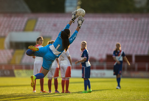Rear view of determined female soccer goalkeeper catching the ball after a free kick on a match at stadium.