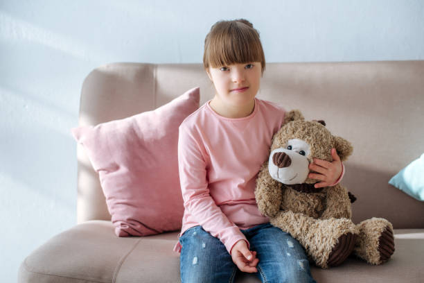 istock Kid with down syndrome sitting on sofa and hugging teddy bear 956331522