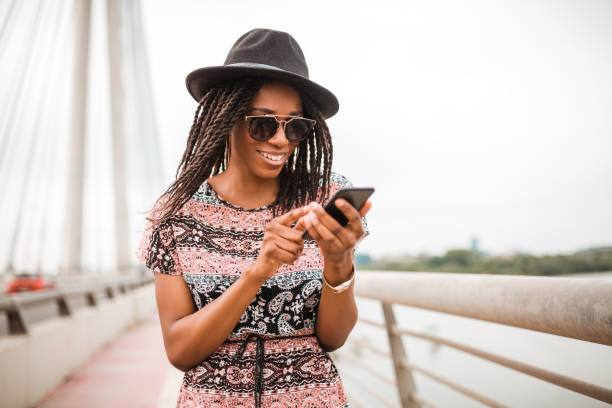 istock Elegant african american woman with hat and sunglasses is browsing internet while standing on a bridge 956334694