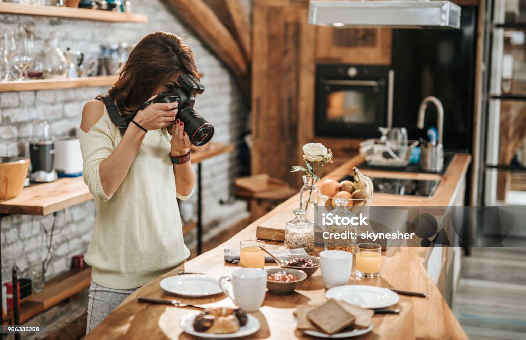 Female photographer taking photos of food at dining table. Young female photographer taking photos of breakfast table in the kitchen. Photographer Stock Photo