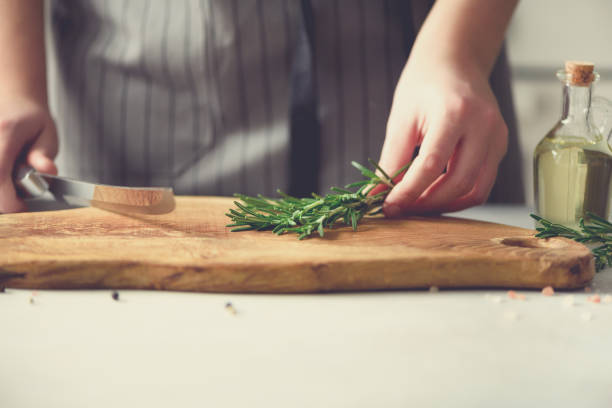 Woman hands cutting fresh green rosemary on wood chopping board in white kitchen, interior. Copy space. Homemade food conceplt, healthy recipe. Take me to work