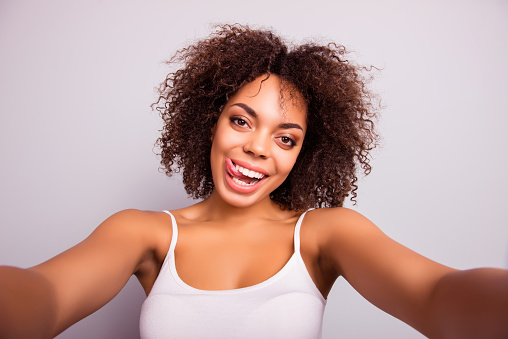 Self portrait of charming, crazy, pretty, sweet, comic, cute attractive girl shooting selfie on front camera with two hands, having beaming smile, showing tongue out, isolated on grey background