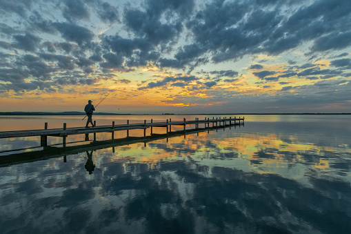 Rear view on silhouette of unrecognizable fisherman walking with fishing rod on jetty under majestic cloudscape at dusk. Location: Lake Steinhuder Meer, Lower Saxony, Germany.