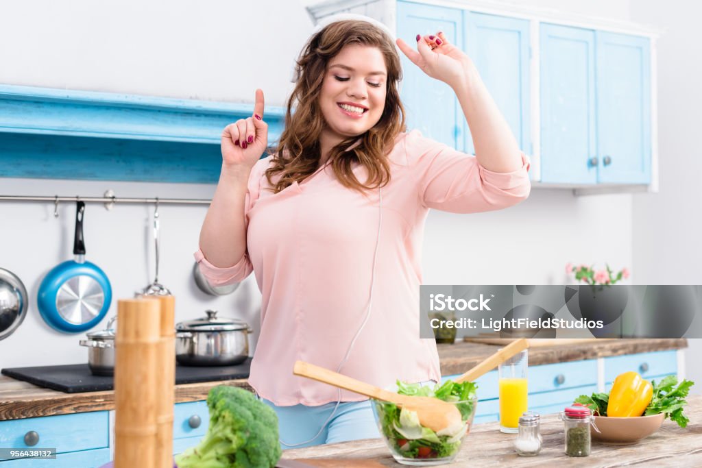 cheerful overweight woman listening music in headphones and dancing at table with fresh vegetables in kitchen at home Overweight Stock Photo