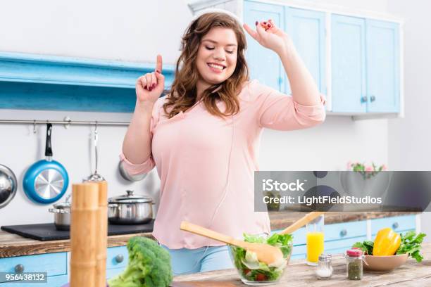 Cheerful Overweight Woman Listening Music In Headphones And Dancing At Table With Fresh Vegetables In Kitchen At Home Stock Photo - Download Image Now