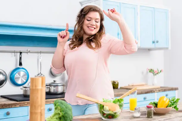 Photo of cheerful overweight woman listening music in headphones and dancing at table with fresh vegetables in kitchen at home
