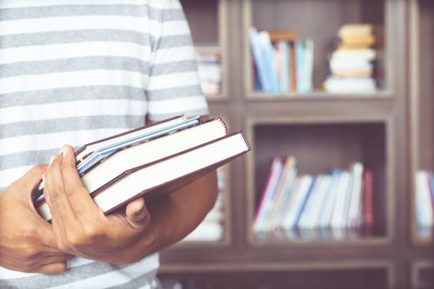 istock close up student young man hand holding books are placed front bookshelf in a library University. love to read education concept. with filter Tones retro vintage warm effect. 956469840