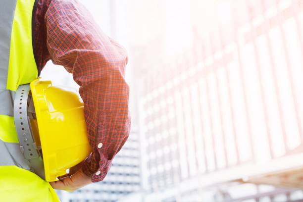 istock Close up backside view of engineering male construction worker stand holding safety yellow helmet and wear reflective clothing for the safety of the work operation. outdoor of building background. 956469996