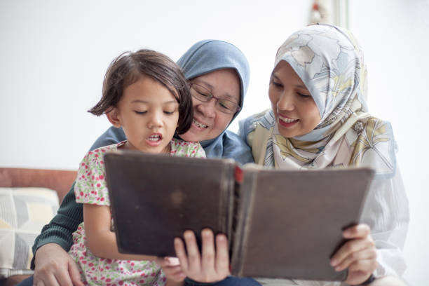 Mother and grandmother reading a book to a child Two Malay woman reading to a young girl using a tablet that looks like a book malay stock pictures, royalty-free photos & images