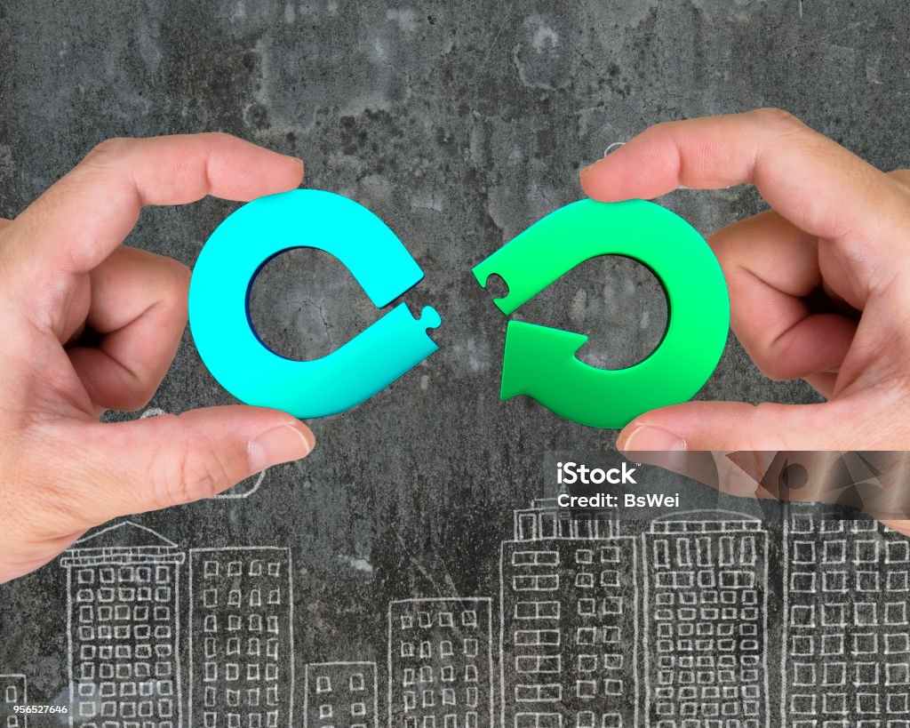 Circular economy concept. Circular economy concept. Two hands assembling arrow infinity recycling symbol of jigsaw puzzle pieces, on city buildings doodles background. Circular Economy Stock Photo