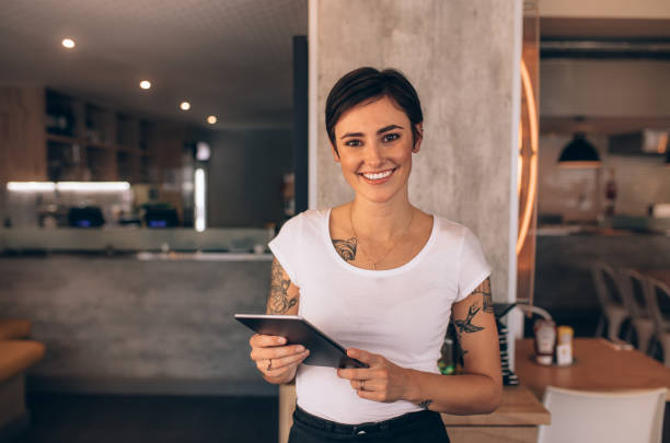 Female restaurant owner with a digital tablet Portrait of a smiling coffee shop owner standing inside her shop. Female restaurant owner standing with a digital tablet. cafe tablet stock pictures, royalty-free photos & images