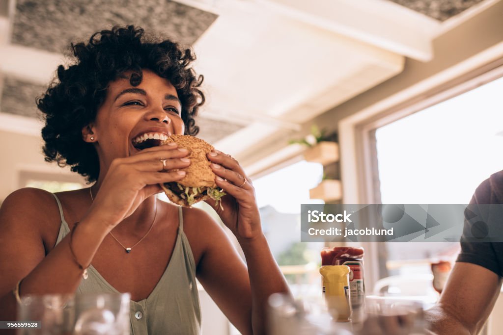 Woman enjoying eating burger at restaurant African woman eating stack burger at restaurant with friends. Happy young woman having junk food at cafe with friends. Eating Stock Photo