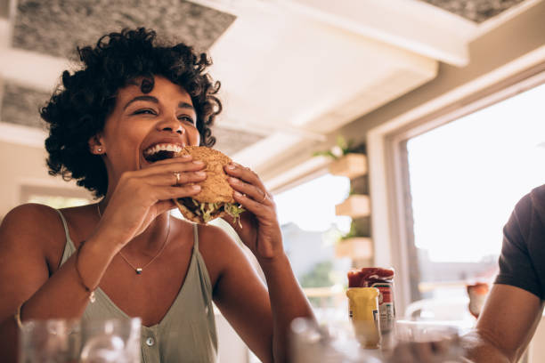 Woman enjoying eating burger at restaurant African woman eating stack burger at restaurant with friends. Happy young woman having junk food at cafe with friends. eating stock pictures, royalty-free photos & images