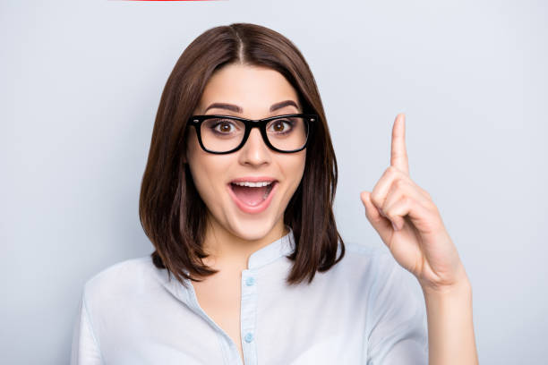 Close up portrait of pretty, charming, glad woman in shirt, eyewear finding an idea, resolution, showing index finger up with open mouth, isolated on grey background Close up portrait of pretty, charming, glad woman in shirt, eyewear finding an idea, resolution, showing index finger up with open mouth, isolated on grey background brain thinking intelligence inspiration stock pictures, royalty-free photos & images