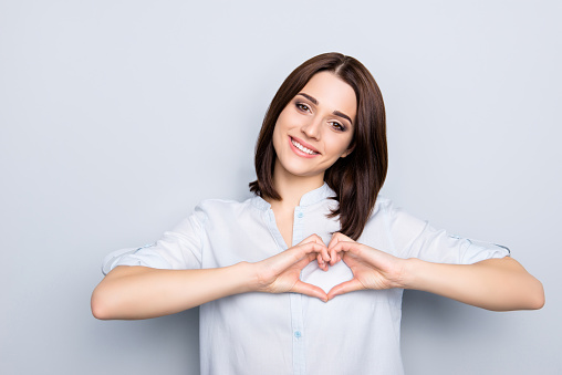Portrait of pretty, charming, stylish, modern, cute, brunette woman with hairstyle in shirt showing love symbol, heart shape with fingers, looking at camera, isolated on grey background
