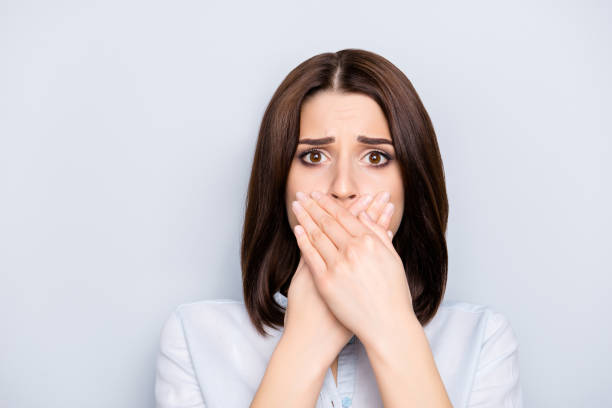 bouchent portrait de séduisantes, joli, mignon, élégant, charmant, femme en chemise fermé sa bouche avec palmes croisées avec une expression inquiète, elle ne peut pas dire n’importe quoi, debout sur fond gris - bad habit fear emotional stress women photos et images de collection