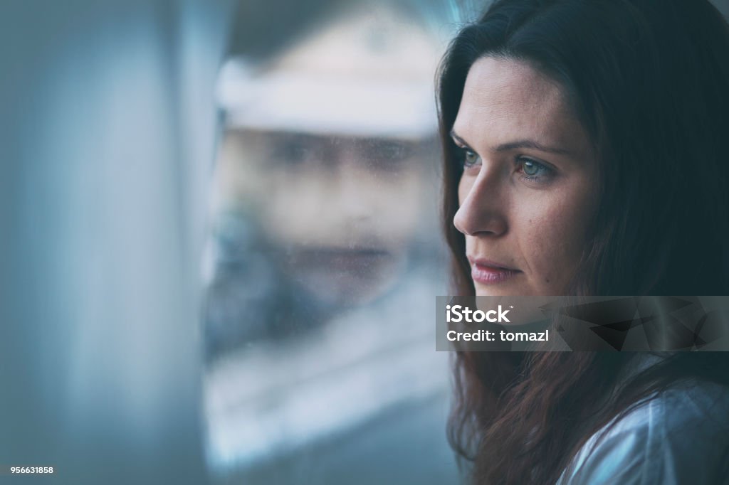Young woman looking through window with reflection Woman with long hair standing by the window so the reflection is visible in the glass. Sadness and anxiety on her face. Women Stock Photo