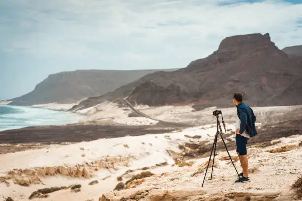 Photo of Photographer with camera in desert admitting unique landscape of sand dunes volcanic cliffs on the Atlantic coast. Baia Das Gatas, near Calhau, Sao Vicente Island Cape Verde