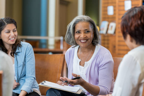 Senior female university professor teaches in casual setting A senior female university professor sits in a circle with her class in the library.  She holds a clipboard as she speaks to the group. career counseling stock pictures, royalty-free photos & images