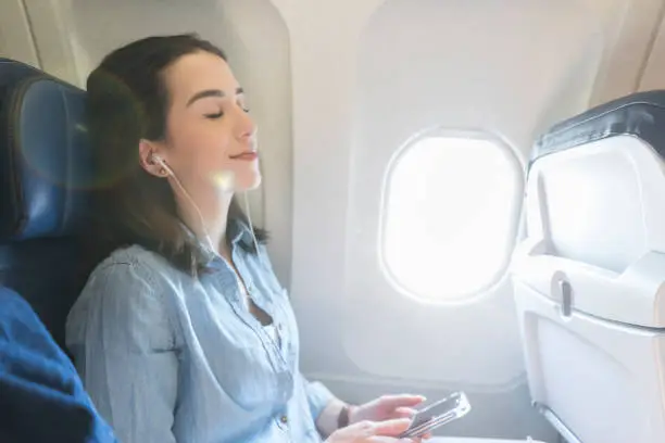 Peaceful young woman closes her eyes while listening to music on a flight. She is sitting next to the window.