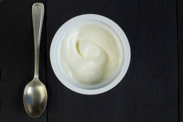 istock Yogurt cup with natural Greek yoghurt in plastic cup with silver spoon closeup on rustic black table background - Top view photo 956781730