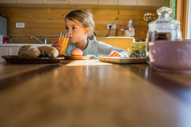 Small pensive girl sitting at dining table and drinking juice during her breakfast time. Copy space.