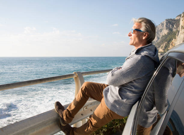 Man leans against car and looks out over Mediterranean Sea He looks off to distant scene with foot up on railing, Liguria finale ligure stock pictures, royalty-free photos & images